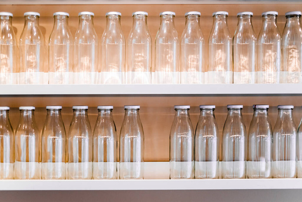 πολλά άδεια γυάλινα μπουκάλια με λευκό καπάκι είναι στο ράφι στη σειρά. μπουκάλι για την αποθήκευση του γάλακτος, χυμό, ποτά και πολλά άλλα. γυάλινο βάζο σε ράφι στην κουζίνα. παραγωγή φιάλης - Φωτογραφία, εικόνα