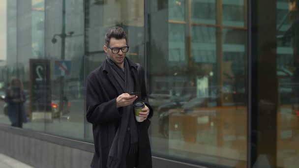 Man in black and eyeglasses with phone walking along the street with coffee - Valokuva, kuva