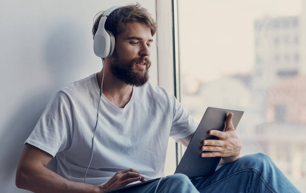 pohledný chlapík ve sluchátkách s laptopem na klíně sedí na parapetu a dívá se z okna - Fotografie, Obrázek