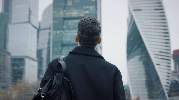 Man in coat walking towards business buildings, slow motion skyscrapers - Fotoğraf, Görsel