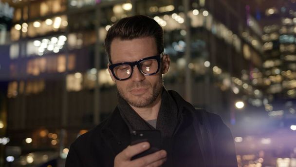 Hombre en gafas con teléfono en el fondo del edificio de negocios en la noche - Foto, Imagen