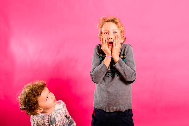 Um menino com gesto espantado e sua irmã mais nova olhá-lo com curiosidade engraçada, isolado em fundo colorido. - Foto, Imagem
