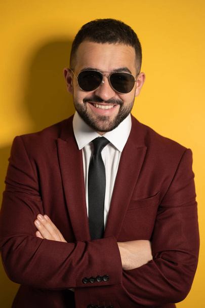Close up portrait of handsome business man in sunglasses and burgundy suit looking at camera with hands folded isolated on yellow background - Fotografie, Obrázek
