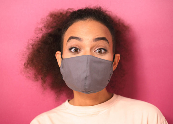 Young looking shocked African American female in light peachy t-shirt in reusable face mask, to prevent others from corona COVID-19 and SARS cov 2 infection isolated on pink background - Foto, Imagen