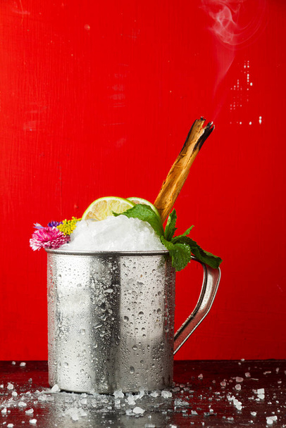Zimny napój w metalowej szklance z zapalonym cynamonem na czarnym stole z czerwonym tłem - Zdjęcie, obraz
