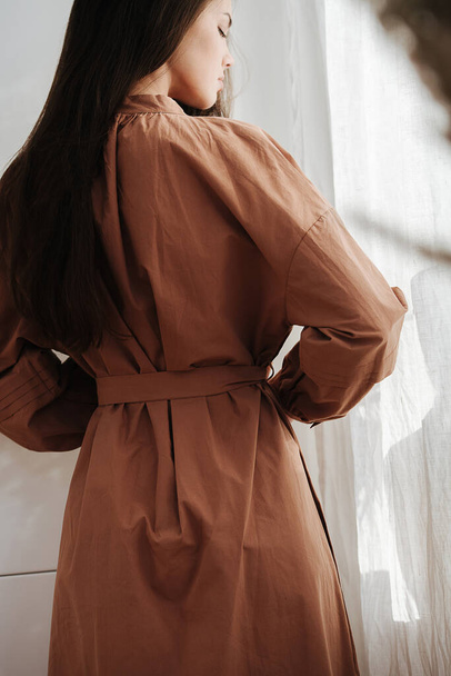 Beautiful woman in a brown dress standing next to a curtain. View from behind. - Foto, imagen