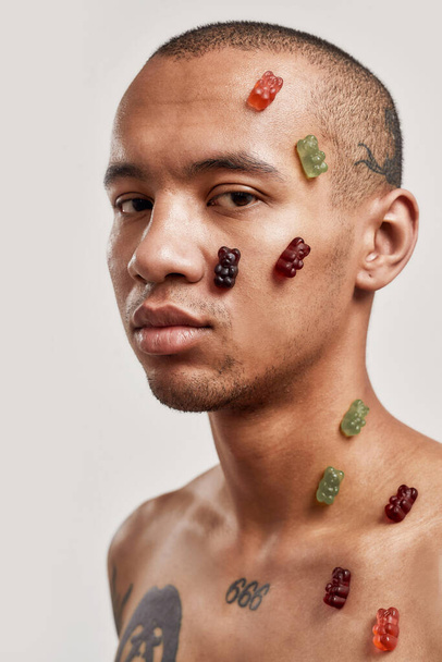 Jelly bears. A dark-skinned tattooed topless male having jellybears stick to his skin while standing sideways looking into a camera - Photo, image
