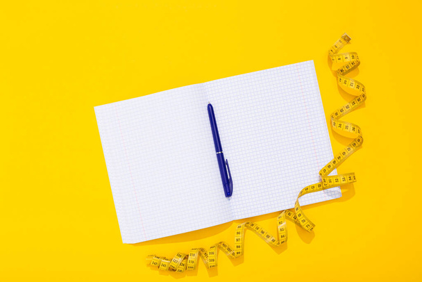 Twisted centimeter tape and clean note book and blue pen with copy space on yellow background. Greeting postcard. Body measurement. Nutrition and fitness workout plan. International Women Day 8 March. - Fotografie, Obrázek