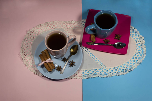 Hay dos tazas de café en una servilleta azul y rosa en platillos. Cerca hay cucharas, palitos de canela y estrellas de anís. - Foto, Imagen