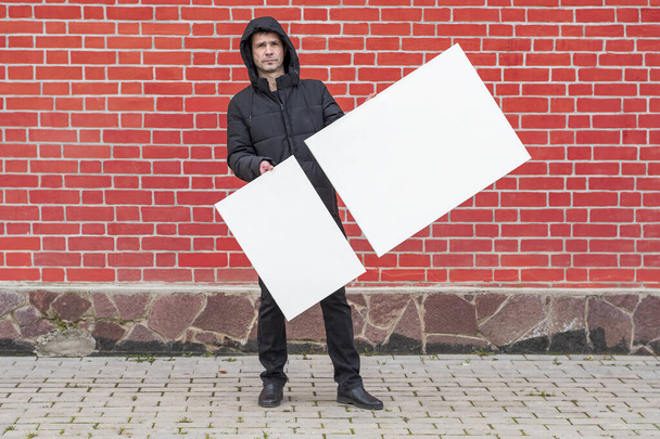 Un jeune homme se tient sur un fond de mur de briques rouges et tient devant lui deux toiles blanches vierges pour la peinture - Photo, image