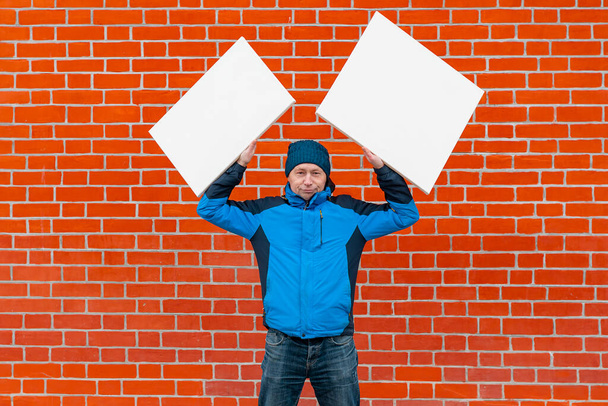 A young man in blue clothes stands against the background of a red brick wall and holds two white blank canvases above his head. - Photo, Image