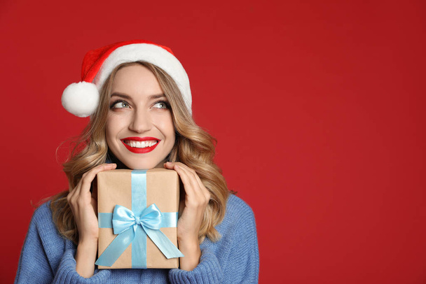 Happy young woman wearing Santa hat with Christmas gift on red background, space for text - Φωτογραφία, εικόνα