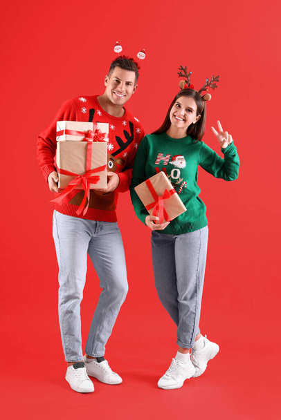 Beautiful happy couple in Christmas headbands and sweaters holding gifts on red background - Photo, Image