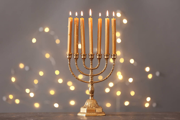 Golden menorah with burning candles on table against grey background and blurred festive lights - Foto, Imagen