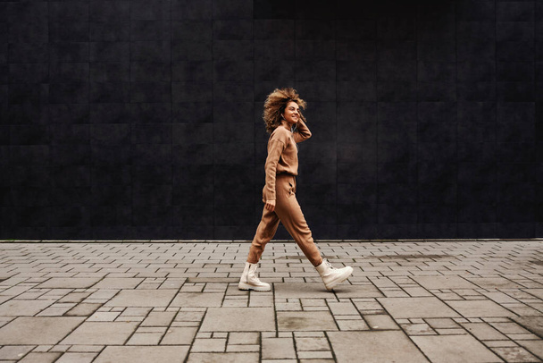 Fiatal, divatos nő göndör hajjal sétál az utcán és zenét hallgat.. - Fotó, kép