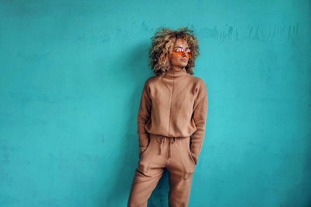 Mulher na moda jovem com cabelo encaracolado de mãos dadas em bolsos e de pé na frente da parede turquesa. - Foto, Imagem