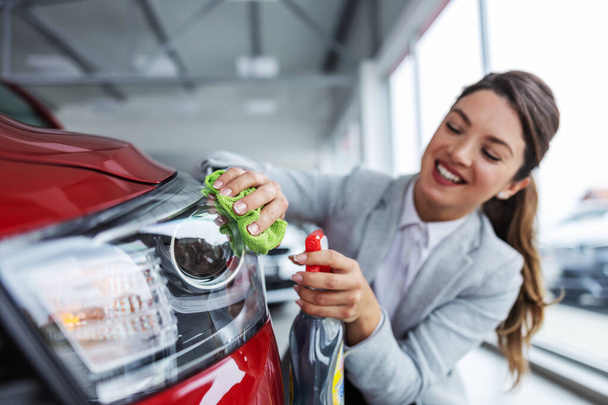 Усміхнена жінка продавець автомобілів у костюмі прив'язує і протирає машину миючим засобом. Інтер'єр салону автомобілів
. - Фото, зображення