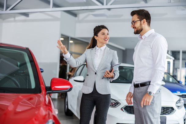 Eine freundlich lächelnde Autoverkäuferin mit Tablet in der Hand spricht mit einem Mann, der ein Auto kaufen will, über die Auto-Spezifikationen. Salon-Innenraum. - Foto, Bild