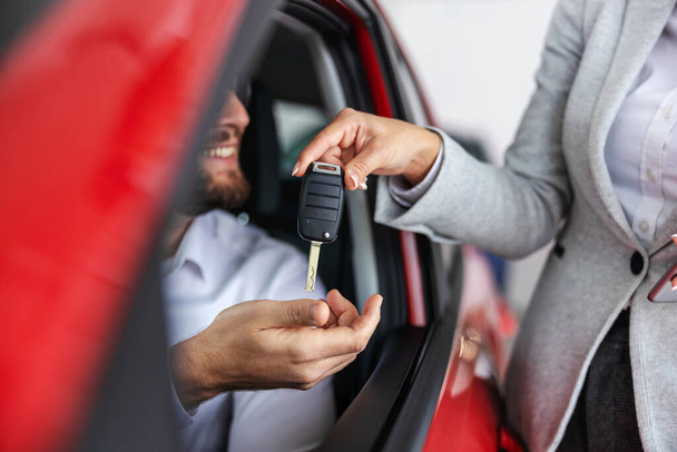 Κοντινό πλάνο του γυναικείου πωλητή αυτοκινήτων που παραδίδει ένα κλειδί αυτοκινήτου σε έναν αγοραστή που κάθεται στο νέο αυτοκίνητο. Σαλόνι αυτοκινήτου εσωτερικό. - Φωτογραφία, εικόνα