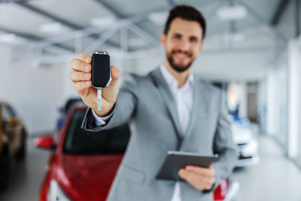 Closeup of car seller holding a key and handing in towards camera while standing in car salon. - 写真・画像