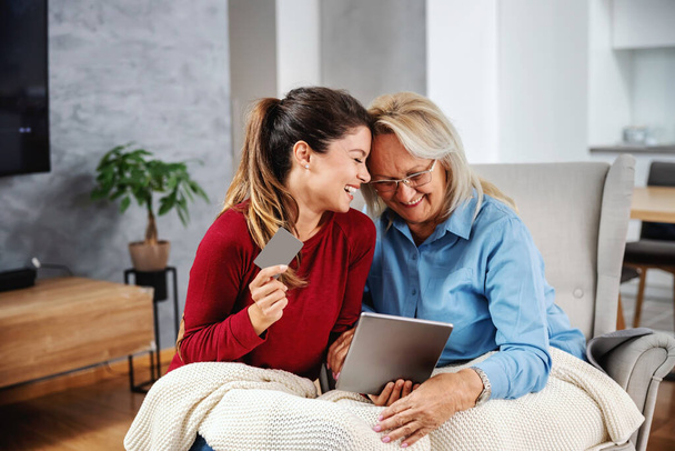 Smiling blond senior woman sitting ina chair and looking at tablet. Her daughter sitting next to her, holding tablet and credit card. They are about to shop online. - Photo, Image