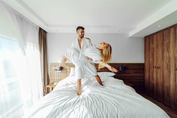 Ein fröhliches Paar mittleren Alters in Bademänteln feiert in einem Hotel seinen Jahrestag. Ein Mann hält eine Frau in seinen Händen. - Foto, Bild