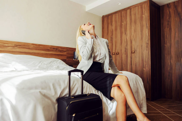 Śmiejąca się kobieta w średnim wieku siedząca na łóżku w pokoju hotelowym i rozmawiająca przez telefon z kimś. Obok niej jest walizka.. - Zdjęcie, obraz
