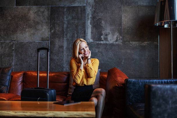 Mujer sonriente de mediana edad en traje sentado en el vestíbulo de un hotel de lujo, teniendo una conversación telefónica y esperando para registrarse en el hotel. - Foto, imagen