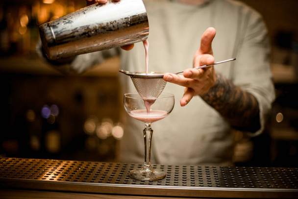 bartender holds sieve over wine glass and gently pours cocktail from shaker. - Фото, изображение