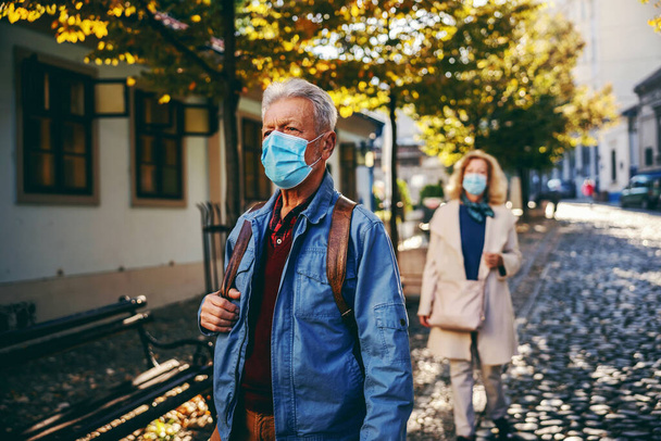 Starszy mężczyzna w masce ochronnej, z plecakiem spacerującym po mieście w słoneczny jesienny dzień. W tle jest też starsza kobieta w masce.. - Zdjęcie, obraz