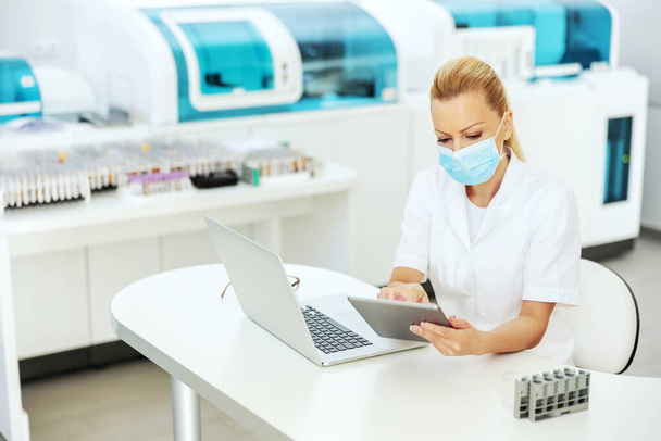 Attractive blond lab assistant in coat with sterile surgical mask on sitting in laboratory, entering test results on tablet. Research for cure for covid 19 concept. - Photo, Image