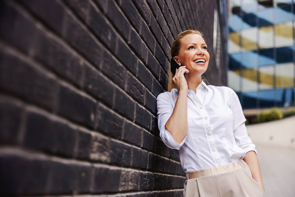 Smiling cheerful fashionable blond businesswoman leaning on the wall outdoors and talking on the phone. - Valokuva, kuva