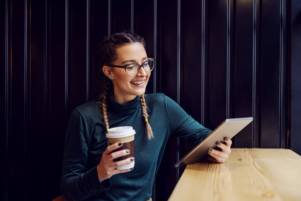 Молодая улыбающаяся женщина сидит в кафе, держа одноразовую чашку с кофе и глядя на планшет. - Фото, изображение