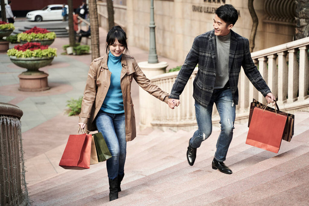 νεαρό ζευγάρι ασιατών που μεταφέρουν τσάντες ψώνια κρατώντας τα χέρια περπάτημα στα σκαλιά, χαρούμενος και χαμογελαστός - Φωτογραφία, εικόνα