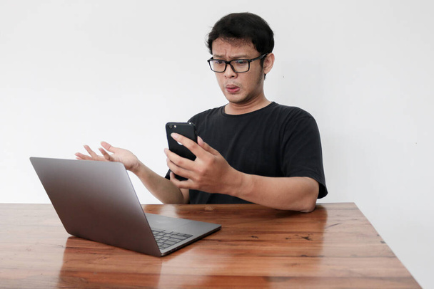 Wow face of Young Asian man shocked and surprised what he see in the smartphone and laptop on the table. Indonesia Man wear black shirt Isolated grey background. - 写真・画像