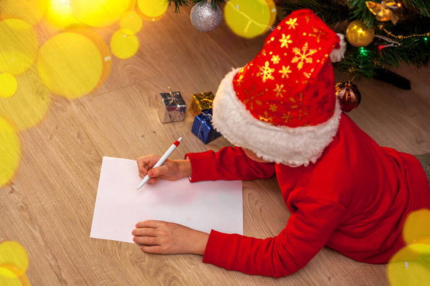 Egy Mikulás sapkás gyerek és egy piros pulcsi fekszik a padlón a karácsonyfa mellett, és kívánságlistát ír. Ünnepi koncepció - Karácsony, újév. A szöveg helye - Fotó, kép