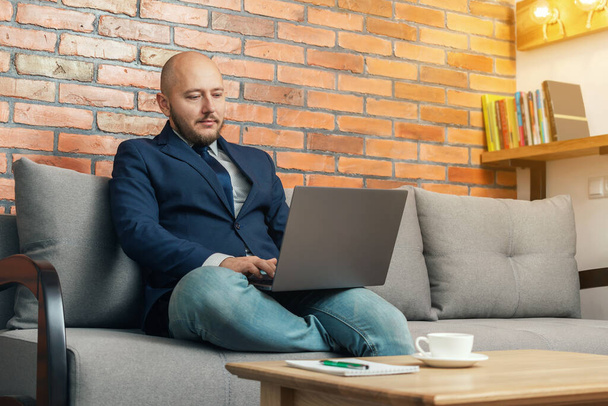 Homem careca barbudo, homem de negócios ou freelancer sentado no sofá e trabalhando no laptop de casa, design moderno loft interior. - Foto, Imagem