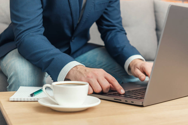 Homme non reconnu, homme d'affaires ou pigiste assis sur un canapé et travaillant à un ordinateur portable sur une table avec une tasse à café, travail de la maison. - Photo, image