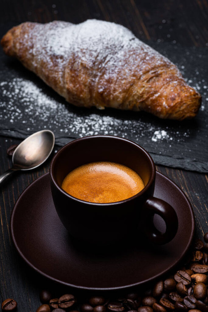 op de donkere rustieke houten tafel, een romige kop Italiaanse koffie op de voorgrond en een croissant bestrooid met poedersuiker. Stilleven - Foto, afbeelding