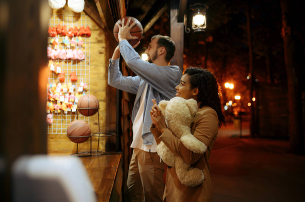 Love couple play in shooting gallery, night amusement park. Man and woman relax outdoors, fairground on background. Family leisures on carousels, entertainment theme - Foto, Imagen