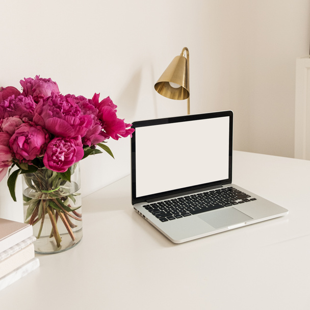 オフィスの机のテーブルワークスペースには、空白のコピースペースがノートパソコンの画面をモック。ピンクの牡丹の花の花束とモダンなスタイリッシュなインテリアデザイン。フロントビューブログ,ソーシャルメディア,ウェブサイトのヒーローテンプレート. - 写真・画像