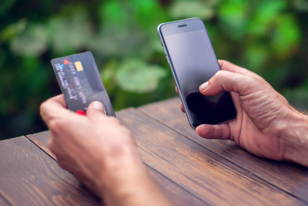 Мужские руки, держащие кредитную карту и используя смартфон для оплаты онлайн на открытом воздухе - Фото, изображение