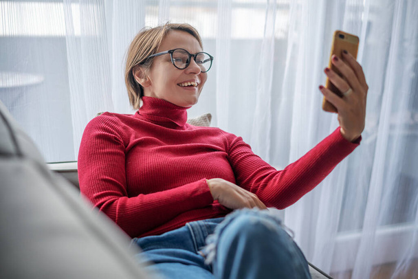 Beautiful smiling happy woman in a red turtleneck at home on a gray sofa lying with a mobile phone in her hands video chatting - Zdjęcie, obraz