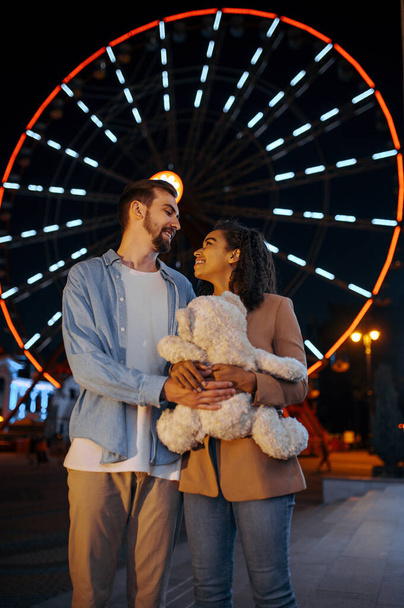 Love couple with soft toy, romantic walking in night amusement park. Man and woman relax outdoors, ferris wheel with lights on background. Family leisures on carousels, entertainment theme - Photo, Image