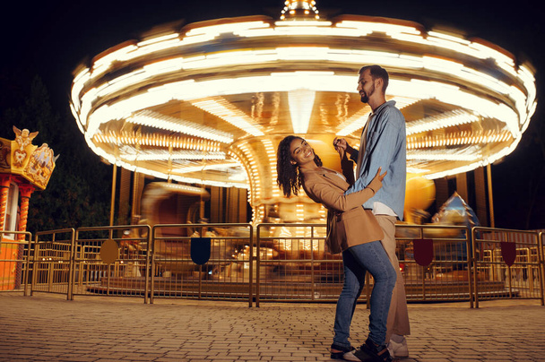 Rakkaus pari tanssia karuselli yöllä huvipuistossa. Mies ja nainen rentoutua ulkona, liikenneympyrä vetovoima valot taustalla. Perheen vapaa-aika kesällä, viihde-teema - Valokuva, kuva