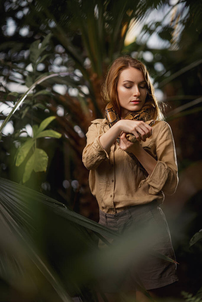 Ένα όμορφο κορίτσι με φυσικό μακιγιάζ και κόκκινα μαλλιά στέκεται στη ζούγκλα ανάμεσα σε εξωτικά φυτά με φίδι. - Φωτογραφία, εικόνα
