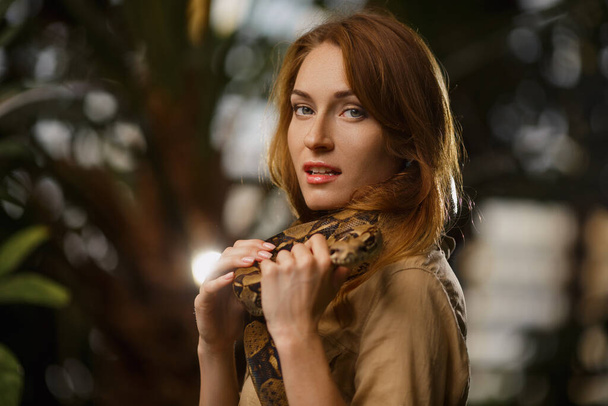 Ein schönes Mädchen mit natürlichem Make-up und roten Haaren steht im Dschungel zwischen exotischen Pflanzen mit Schlange. - Foto, Bild