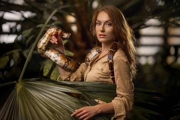 Kaunis tyttö, jolla on luonnollinen meikki ja punaiset hiukset seisoo viidakossa eksoottisten kasvien joukossa käärme. - Valokuva, kuva