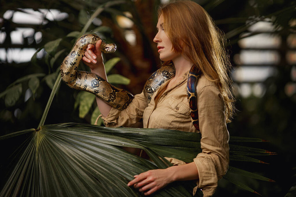 Une belle fille au maquillage naturel et aux cheveux roux se tient dans la jungle parmi les plantes exotiques avec serpent. - Photo, image