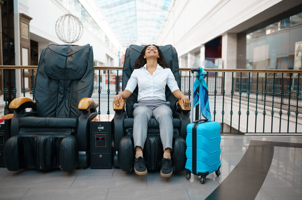 Woman with suitcase relax in massage chair, airport waiting hall. Female person with luggage, international air gate, passenger with bag. Girl with baggage in departure terminal - Photo, Image